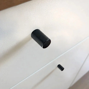 Gros plan de deux boutons Remo Black Deluxe de yessential.fr sur un boîtier blanc, leur design texturé projetant des ombres subtiles. L’arrière-plan flou attire l’attention sur ces détails de boutons d’armoire modernes.