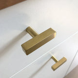 Un gros plan de deux boutons Vasco Gold modernes de yessential.fr, avec une finition dorée luxueuse et une finition brossée, montés verticalement sur une façade de tiroir blanche.