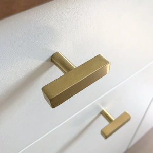 Un gros plan de deux boutons Vasco Gold modernes de yessential.fr, avec une finition dorée luxueuse et une finition brossée, montés verticalement sur une façade de tiroir blanche.