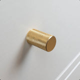 Un gros plan du bouton Remo Gold Deluxe, un cylindre en laiton texturé, monté sur une façade d'armoire ou de tiroir blanche.