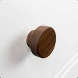 Le bouton Arriero Walnut de yessential.fr est élégamment monté sur une façade d'armoire ou de tiroir structurée blanche, avec un subtil bord surélevé visible à l'arrière-plan.