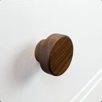 Le bouton Arriero Walnut de yessential.fr est élégamment monté sur une façade d'armoire ou de tiroir structurée blanche, avec un subtil bord surélevé visible à l'arrière-plan.