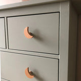 Gros plan de la commode Bambino Moon de yessential.fr, avec tiroirs gris clair avec boutons en bois en forme de lune. Avec sa finition mate lisse et son design moderne, c'est un choix élégant pour toute chambre de bébé.