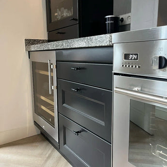 Cuisine moderne avec armoires noires avec le bouton Cordero Black de yessential.fr en noir mat, refroidisseur à vin encastré, four en acier inoxydable avec 8:34, comptoirs en granit moucheté, verrerie dans l'armoire supérieure et cafetière sur le comptoir.