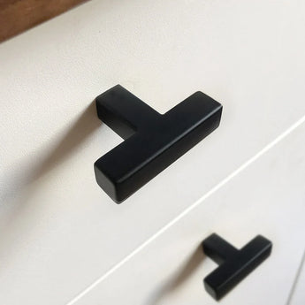 Gros plan d'une armoire blanche avec un bouton Vigio Black moderne noir mat de yessential.fr. Ces poignées en forme de T apportent une touche élégante lorsqu'elles sont montées horizontalement sur deux tiroirs.