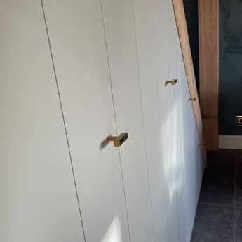 Une rangée d'armoires encastrées blanches avec une luxueuse finition dorée et un bouton Vasco Gold de yessential.fr est installée sous un escalier en bois, à côté d'un mur bleu foncé et d'un sol carrelé gris.
