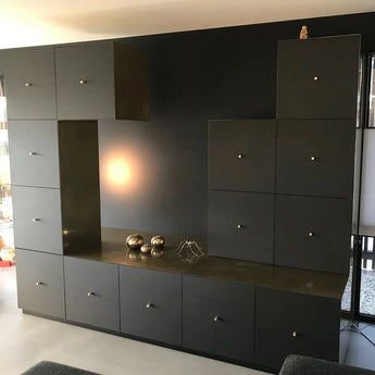 Une unité murale noire moderne avec plusieurs armoires et tiroirs est équipée de luxueux boutons Remo Gold Deluxe de yessential.fr. Des sphères métalliques, une sculpture géométrique sur le comptoir et une statue de hibou dans le coin supérieur droit complètent le look.