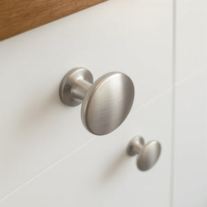 Gros plan de deux boutons de meuble Pedro en acier inoxydable 10x de yessential.fr en acier inoxydable brossé sur des portes d'armoire blanches, avec un bord en bois visible en haut.