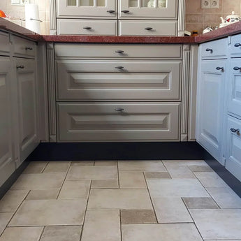 Une cuisine avec des armoires gris clair, des poignées Alvaro Anthracite de yessential.fr et un comptoir rouge. Les carreaux de sol beiges et marron forment un motif. De petites pièces décoratives et un rouleau d'essuie-tout décorent le comptoir.