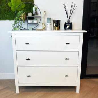 Une commode blanche à deux niveaux et les boutons Remo Black de yessential.fr avec commode sur parquet. Les vases, les plantes, les bougies et un miracle à la surface du mur.