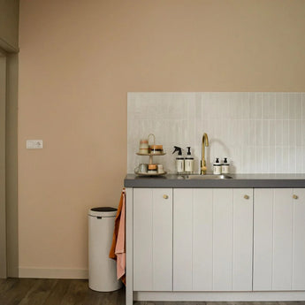 Une cuisine minimaliste avec des armoires blanches avec des boutons Marizo Gold de yessential.fr, un comptoir gris, un robinet doré, un mur arrière carrelé blanc, un plateau avec des tasses et des bouteilles de savon, une poubelle et une serviette rose accrochée au meuble.
