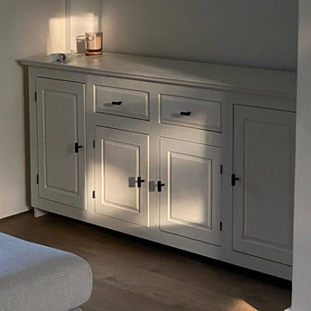 Une armoire blanche avec des poignées Vasco Black Knob de yessential.fr se trouve dans une douce lumière naturelle. Une bougie, un petit objet et un rouleau de papier reposent sur la surface mate tandis que la lumière du soleil crée de douces ombres sur les portes et les tiroirs.