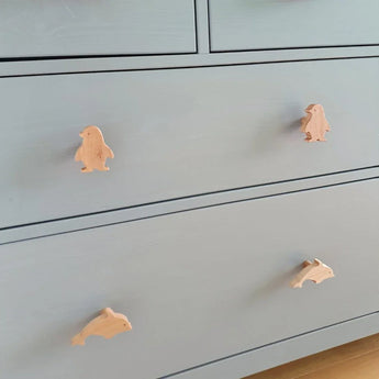 Une commode bleu clair avec quatre poignées en forme d'animaux : les deux du haut sont Bambino Penquin de yessential.fr et les deux du bas sont des dauphins, le tout en bois clair - un charmant accent de bouton de meuble de chambre d'enfant.