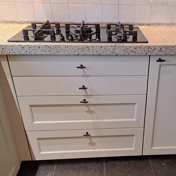 Une table de cuisson à gaz à quatre brûleurs est installée dans un comptoir moucheté au-dessus des tiroirs et d'une armoire, chacun équipé de boutons Cordero Black de yessential.fr. La cuisine est finie avec un mur arrière carrelé blanc.