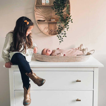 Une jeune fille avec une épingle à cheveux et des bottes dorées est assise sur une commode blanche avec les boutons Vasco Gold de yessential.fr et sourit à un bébé dans un panier tressé. Une étagère murale avec des plantes et un décor au-dessus ajoute de la chaleur à la scène chaleureuse.