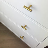 Trois tiroirs blancs avec des boutons modernes Remo Gold de yessential.fr, avec un design horizontal en forme de T, sont présentés sur un plancher en bois clair. L'image montre la partie supérieure des tiroirs vue sous un angle.
