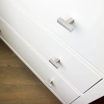 Gros plan d'une commode blanche avec trois tiroirs, chacun équipé d'un bouton Vigio en acier inoxydable de yessential.fr, avec un design élégant en forme de T argenté sur un plancher en bois clair.