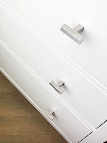 Gros plan d'une commode blanche avec trois tiroirs, chacun équipé d'un bouton Vigio en acier inoxydable de yessential.fr, avec un design élégant en forme de T argenté sur un plancher en bois clair.