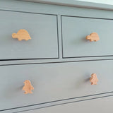 La table à langer Bambino Dino de yessential.fr est gris clair et comporte quatre boutons d'animaux en bois ludiques - deux tortues en haut et deux pingouins en bas - ce qui en fait le complément joyeux parfait à toute chambre d'enfant.