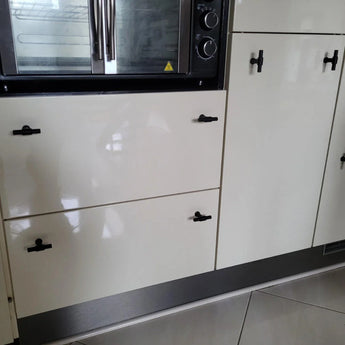 Gros plan d'armoires et de tiroirs de cuisine blanc brillant avec poignées yessential.fr Cordero Black Knob au fini noir mat, sous un four encastré avec finition en acier inoxydable. Il y a de grands carreaux de couleur claire au sol.