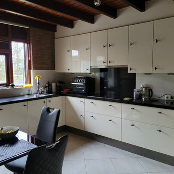 Cuisine moderne avec armoires de couleur crème, comptoirs noir mat et appareils électroménagers intégrés. Les armoires sont équipées de boutons Cordero Black de yessential.fr. La lumière naturelle entre, avec un ensemble de salle à manger noir et des ustensiles de cuisine élégants sur le comptoir.