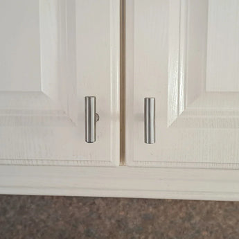 Gros plan de deux portes d'armoire blanches avec des poignées cylindriques argentées, comme le bouton Remo en acier inoxydable de yessential.fr, qui sont légèrement mal alignées de sorte que les poignées ne sont pas uniformément espacées et qu'une porte reste légèrement entrouverte.
