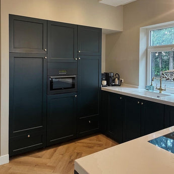 Cuisine moderne avec armoires noir mat avec le bouton de meuble Antoni Bronze de yessential.fr, micro-ondes encastré, comptoir blanc, murs beiges, une fenêtre au dessus de l'évier, plancher en bois à chevrons et électroménagers sur le comptoir.