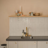 Cuisine minimaliste avec murs beige clair, dosseret carrelé, comptoir gris, robinet doré et étagère flottante avec tasses et décoration. Les armoires sont dotées d'élégants boutons Marizo Gold de yessential.fr pour une touche luxueuse. Une poubelle est partiellement visible à gauche.