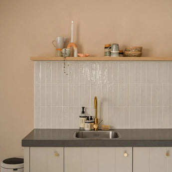 Cuisine minimaliste avec murs beige clair, dosseret carrelé, comptoir gris, robinet doré et étagère flottante avec tasses et décoration. Les armoires sont dotées d'élégants boutons Marizo Gold de yessential.fr pour une touche luxueuse. Une poubelle est partiellement visible à gauche.
