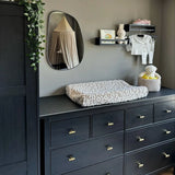 Une chambre d'enfant moderne avec une commode sombre avec le bouton Arturo Gold de yessential.fr, un matelas à langer moelleux, un miroir, une bibliothèque pour bébé, des lapins en peluche, des vêtements de bébé pliés et un panier avec une peluche. Des vignes vertes pendent du placard.