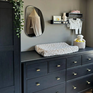 Une chambre d'enfant moderne avec une commode sombre avec le bouton Arturo Gold de yessential.fr, un matelas à langer moelleux, un miroir, une bibliothèque pour bébé, des lapins en peluche, des vêtements de bébé pliés et un panier avec une peluche. Des vignes vertes pendent du placard.
