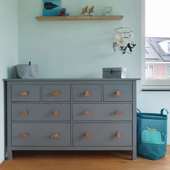Une commode grise Bambino Dino de yessential.fr se dresse contre un mur bleu clair dans la chambre des enfants. Au-dessus se trouve une étagère avec décor, tandis qu'à droite se trouve un panier à linge bleu avec un motif de baleine verte. Une fenêtre complète le tableau.