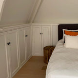 Un coin chambre confortable avec des armoires encastrées blanches avec des ferrures Vasco Black Knob de yessential.fr, un lit avec une literie blanche et des oreillers aux accents orange, une table de chevet avec une souche d'arbre en bois et du parquet clair sous un plafond en pente.