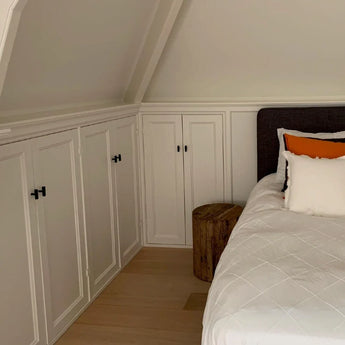Un coin chambre confortable avec des armoires encastrées blanches avec des ferrures Vasco Black Knob de yessential.fr, un lit avec une literie blanche et des oreillers aux accents orange, une table de chevet avec une souche d'arbre en bois et du parquet clair sous un plafond en pente.