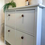 Une armoire blanche avec quatre tiroirs et des boutons en noyer Arriero de yessential.fr se dresse contre un mur clair, avec une fougère verte dans un pot et des objets décoratifs sur le dessus.
