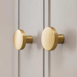 Deux boutons ronds Marizo Gold de yessential.fr sont installés sur les portes d'armoires gris clair, vues en gros plan. Ces boutons élégants ont une finition dorée brossée lisse et légèrement réfléchissante.