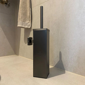 La brosse de toilette Alessino Gunmetal de yessential.fr, avec un support carré, se pose sur le sol clair de la salle de bain avec une serviette beige et avec une touche élégante d'accessoires de salle de bain et bronze.