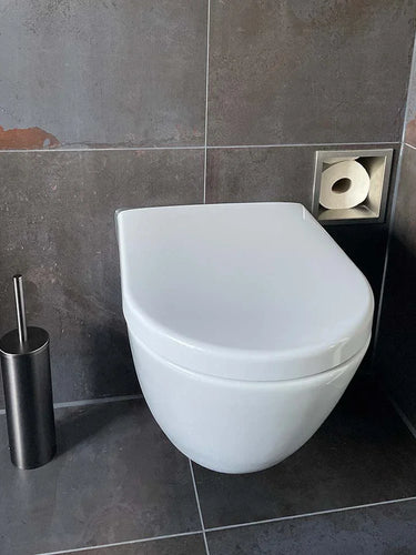 Des toilettes murales blanches modernes avec un couvercle fermé, à côté de l'ensemble WC Tygo Gunmetal (sans perçage) de yessential.fr et d'un porte-papier toilette carré encastré dans une salle de bains aux murs et au sol carrelés gris foncé.
