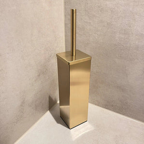 La brosse de toilette Velasco Gold de yessential.fr, autoportante avec support, se trouve dans le coin d'une salle de bain aux murs et au sol carrelés beige.