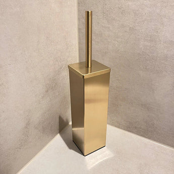 La brosse de toilette Velasco Gold de yessential.fr, autoportante avec support, se trouve dans le coin d'une salle de bain aux murs et au sol carrelés beige.