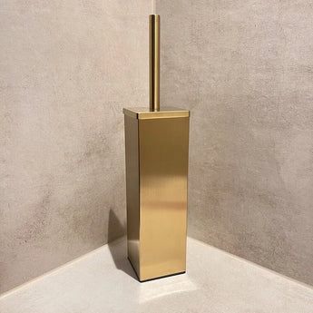 Une brosse de toilette Velasco Gold de yessential.fr, autoportante avec support, se trouve dans le coin d'une salle de bains carrelée beige.