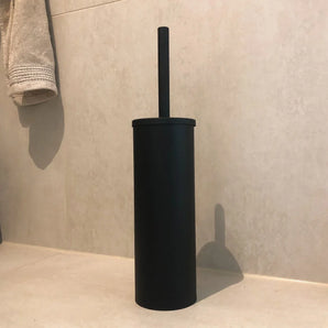 La brosse de toilette autoportante Bruno Black de yessential.fr avec support cylindrique se dresse sur le sol lumineux d'une salle de bain contre un mur carrelé, avec une serviette pliée à proximité.