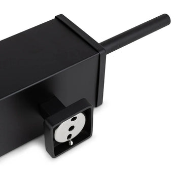 Gros plan sur la multiprise Alessino Black de yessential.fr, avec une prise européenne et un cordon noir, placée à côté d'une brosse WC noire mate pour un style de salle de bain épuré et moderne.
