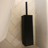 La brosse de toilette Alessino Black avec support de yessential.fr se dresse sur un sol carrelé de salle de bain avec des carreaux de couleur crème en diagonale.