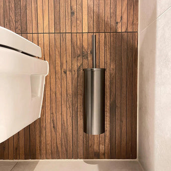 L'ensemble WC Pacho Gunmetal de yessential.fr, avec sa finition métallique élégante, est fixé au mur à côté d'une toilette blanche et apporte un style moderne et des accessoires de salle de bain luxueux à cette salle de bain minimaliste.