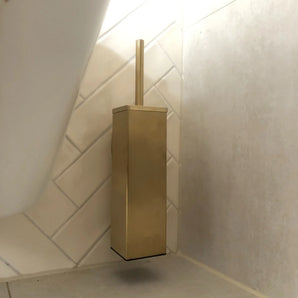 Le porte-brosse de toilette Alessino Gold de yessential.fr, équipé d'une poignée assortie, est monté dans le coin près du sol contre des carreaux blancs et beiges élégants et ajoute une touche d'élégance à votre salle de bain.