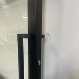 Gros plan d'une poignée de porte en métal noir sur une porte en verre, avec un adhésif pour butée de porte yessential.fr 6x ⌀2 cm fixé au mur blanc à proximité pour empêcher la poignée de heurter le mur.