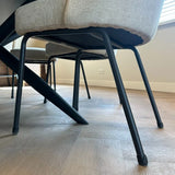 Gros plan de deux chaises modernes avec pieds en métal noir équipées de protège-pieds de chaise anti-rayures yessential.fr Paco Set Ø 17-21 mm, coussins légers, sur un parquet près d'une table. Prise murale et fenêtre avec stores en arrière-plan.