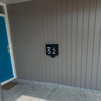 Une boîte aux lettres noire moderne avec des chiffres Animo en acier inoxydable de 15 cm (sans perçage) de yessential.fr avec le texte « 52 » est montée sur un mur lambrissé gris clair à côté d'une porte bleu vif et d'un paillasson dans une entrée carrelée.