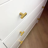 Gros plan d'une commode blanche avec des boutons Vigio Gold de yessential.fr sur trois tiroirs, chacun décoré de poignées dorées modernes en forme de T, sur un parquet marron clair.